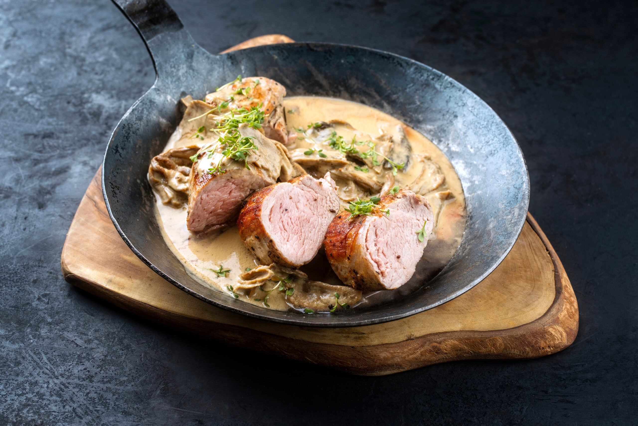 Traditional barbecue Iberian pork filet medaillons in cream sauce with mushrooms and herbs offered as close-up in a rustic wrought-iron pan on a wooden board